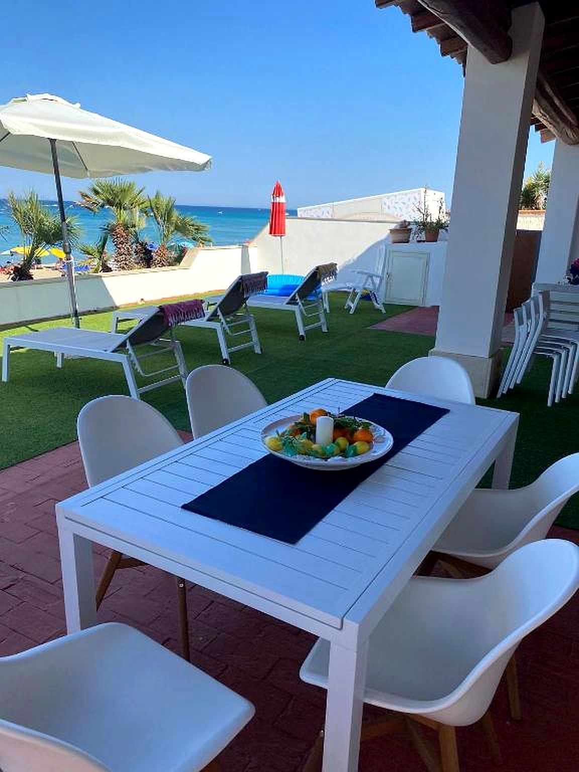 Zona pranzo all'aperto con tavolo bianco, sedie e vista mare.