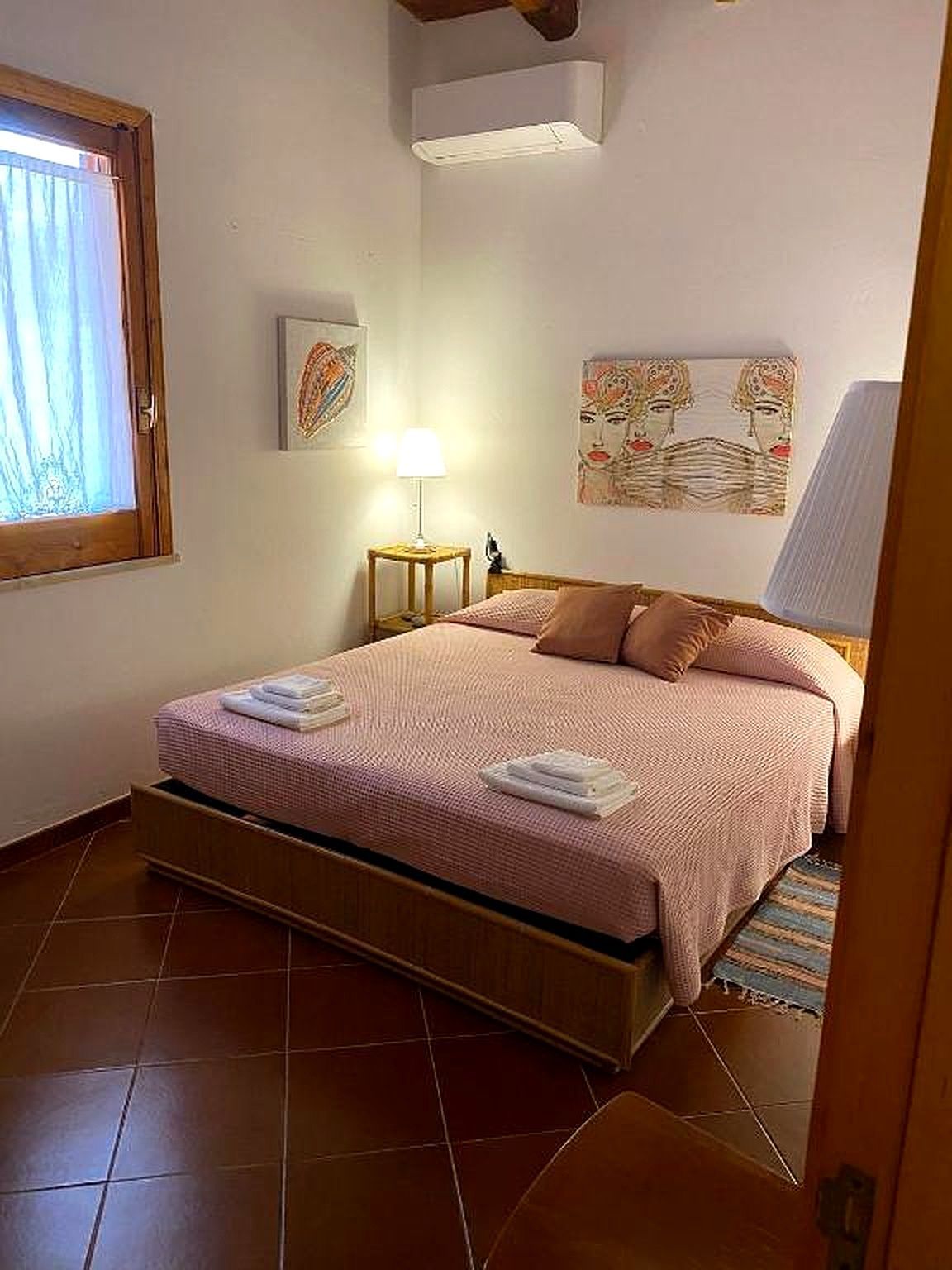 Camera da letto con un letto, due cuscini e una coperta rosa, vicino a una finestra e a un'opera d'arte.