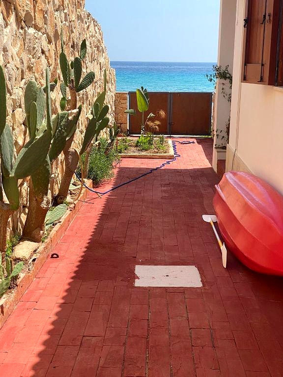 Sentiero di mattoni rossi che conduce al mare. Cactus sul muro di pietra, barca rossa, acqua blu. Giornata di sole splendente.