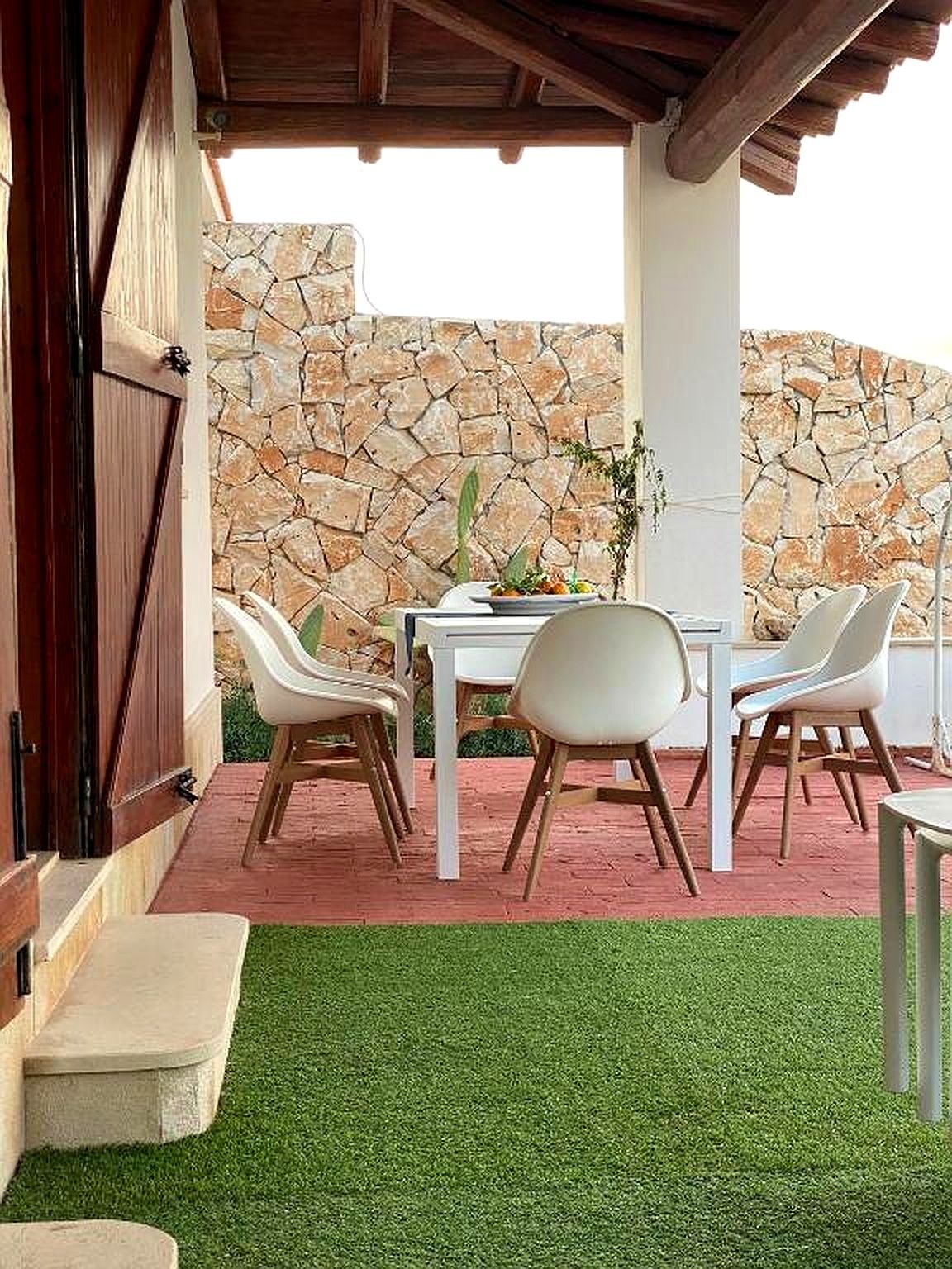 Zona pranzo all'aperto con tavolo bianco, sedie sul patio, muro in pietra e prato verde.