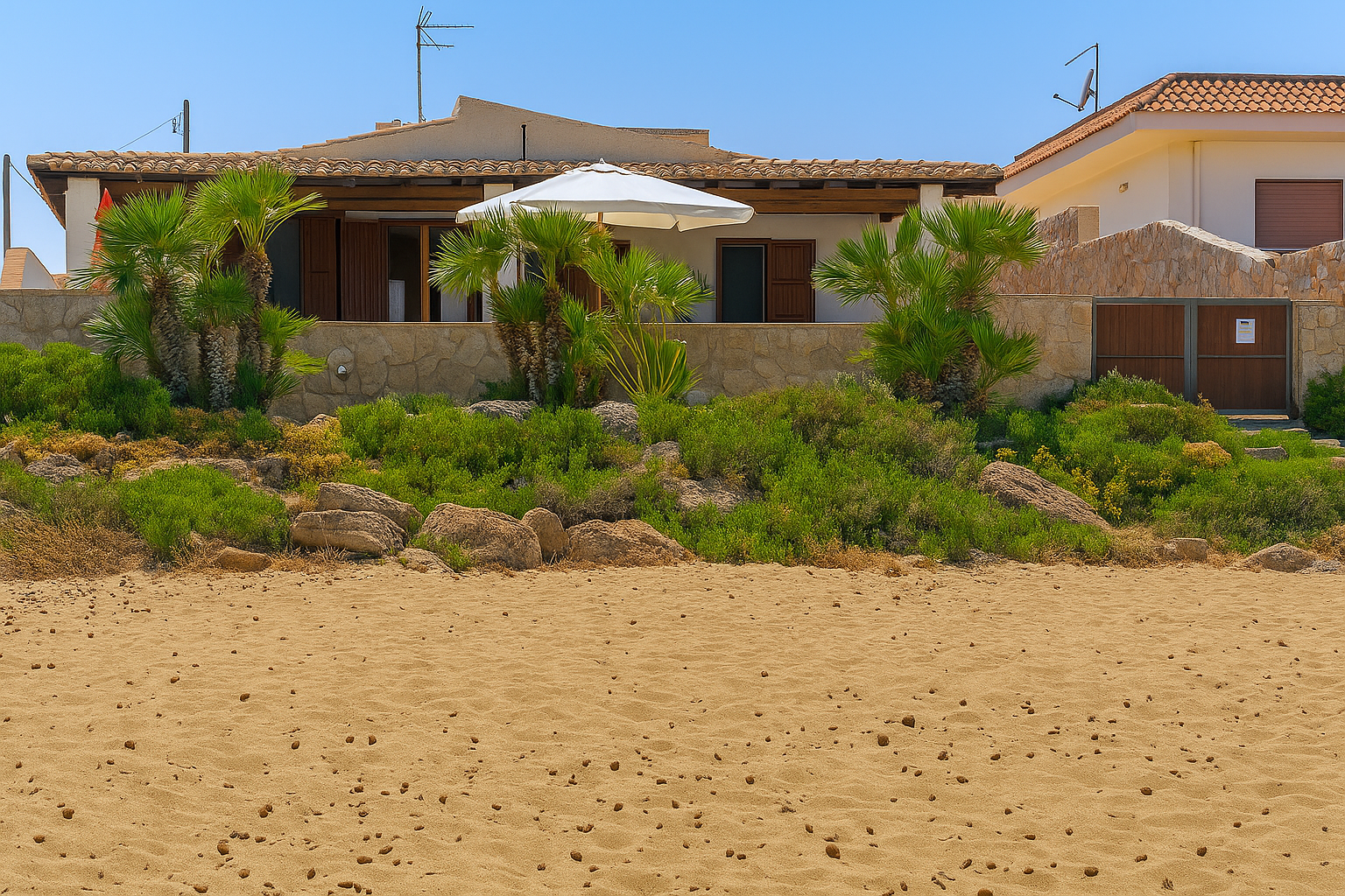 Casa sulla spiaggia con facciata in pietra, vegetazione e ombrellone, sotto un cielo azzurro e terso.