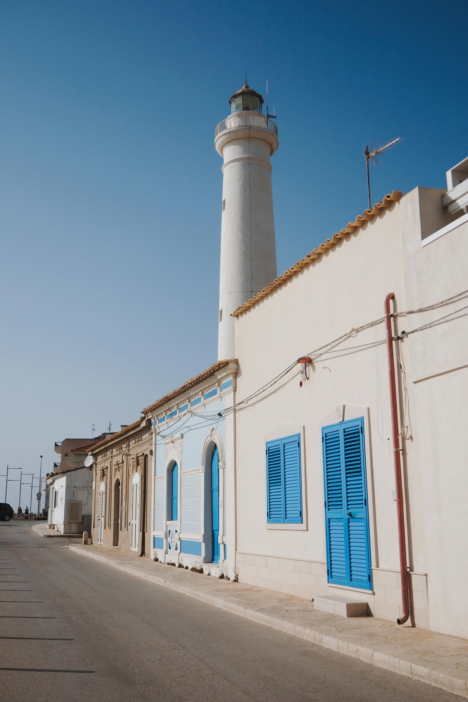 Un faro bianco svetta su edifici chiari con persiane e finiture blu; una strada in una giornata di sole.
