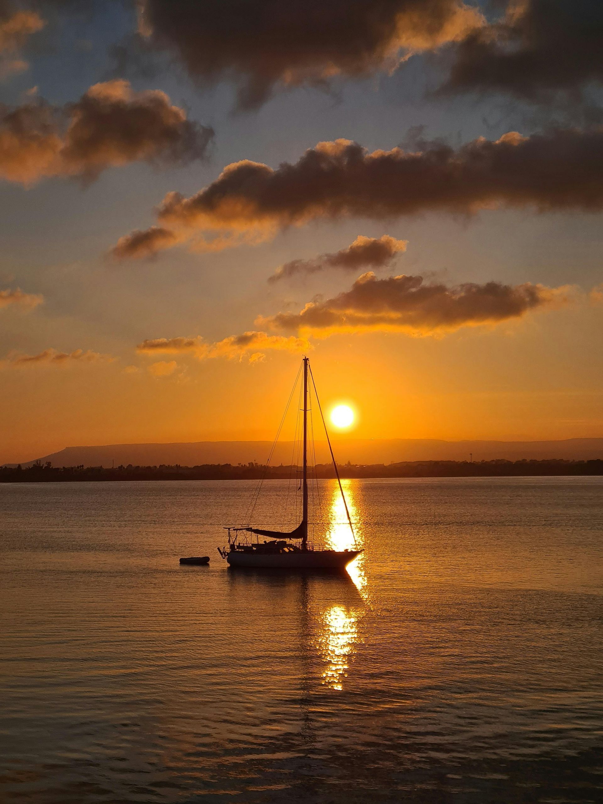 Barca a vela sulle acque calme al tramonto, che riflette il sole arancione brillante e le nuvole.