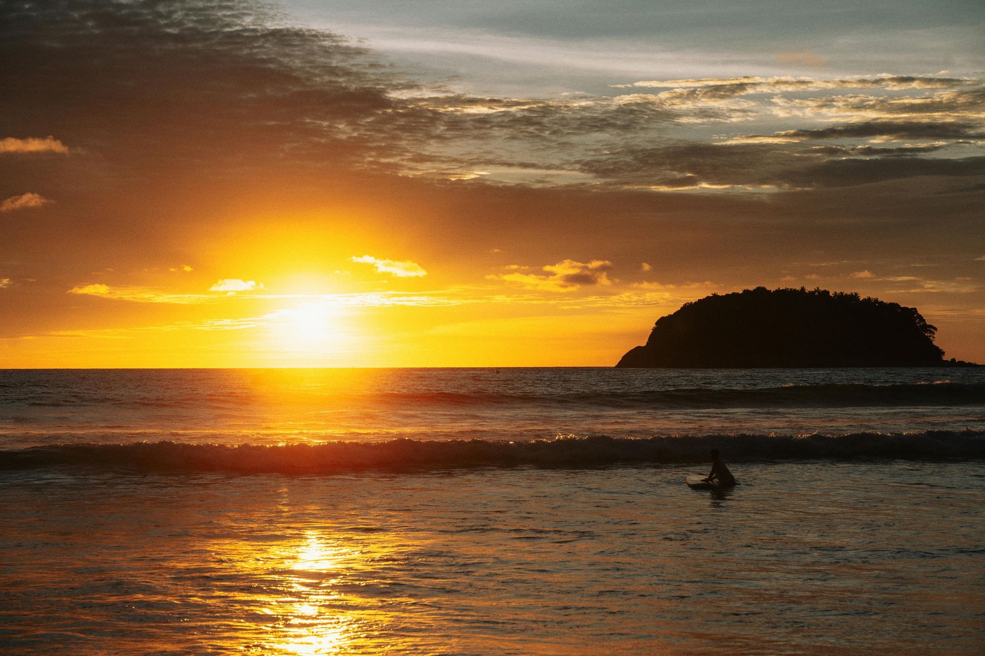 Tramonto sull'oceano con un'isola in silhouette e una persona in acqua. Dominano le tonalità arancioni e dorate.