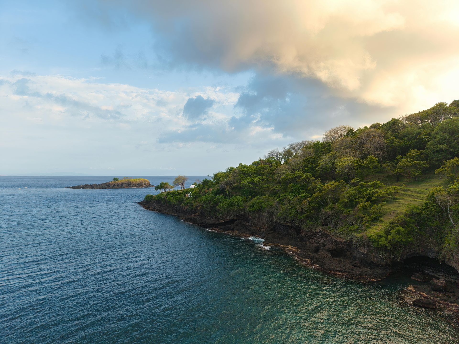 Vista costiera: acqua dell'oceano, costa rocciosa con vegetazione verde, cielo nuvoloso.