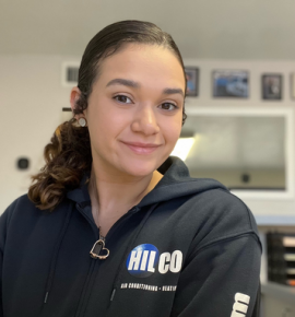 Woman in a black hoodie with logo, smiles at the camera. Dark curly hair, indoor setting.