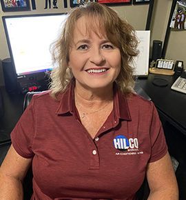 Woman in a maroon shirt with logo smiles at the camera, seated at a desk with a computer.