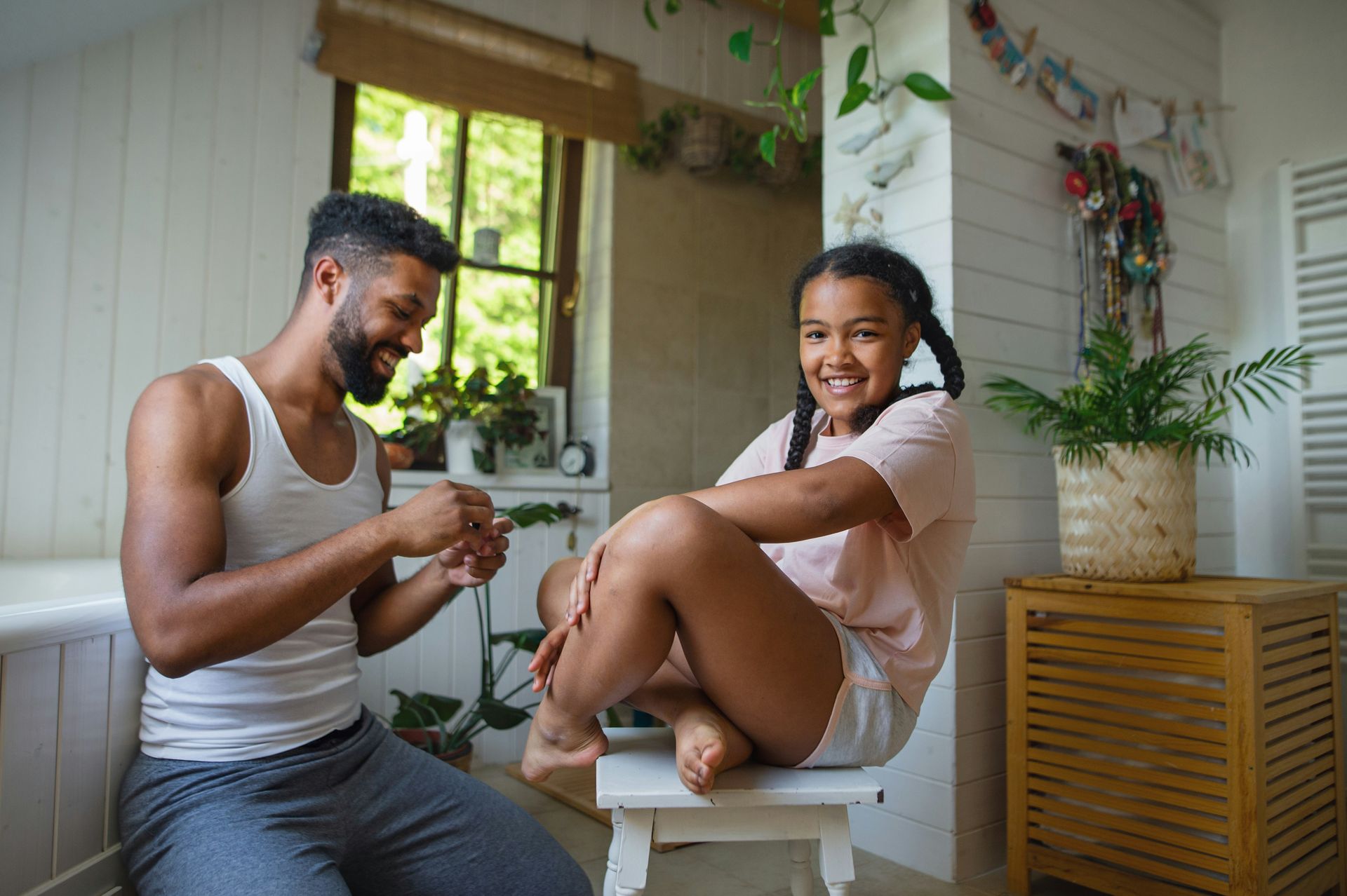 Man painting girl's toenails in a bathroom. She smiles, sitting on a stool. Natural light shines.
