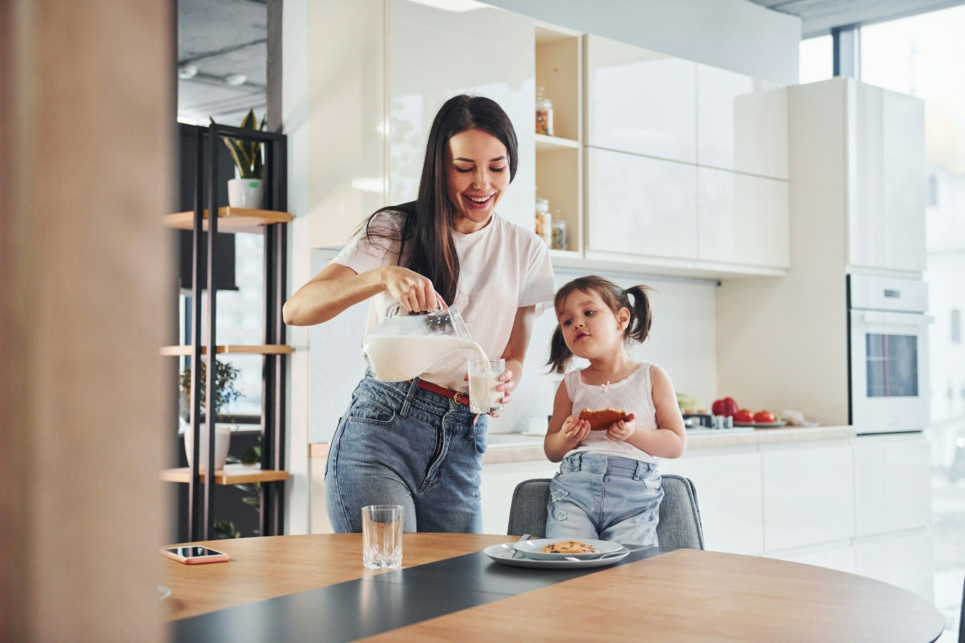 Woman pouring milk for a child in a modern kitchen. Both are smiling.