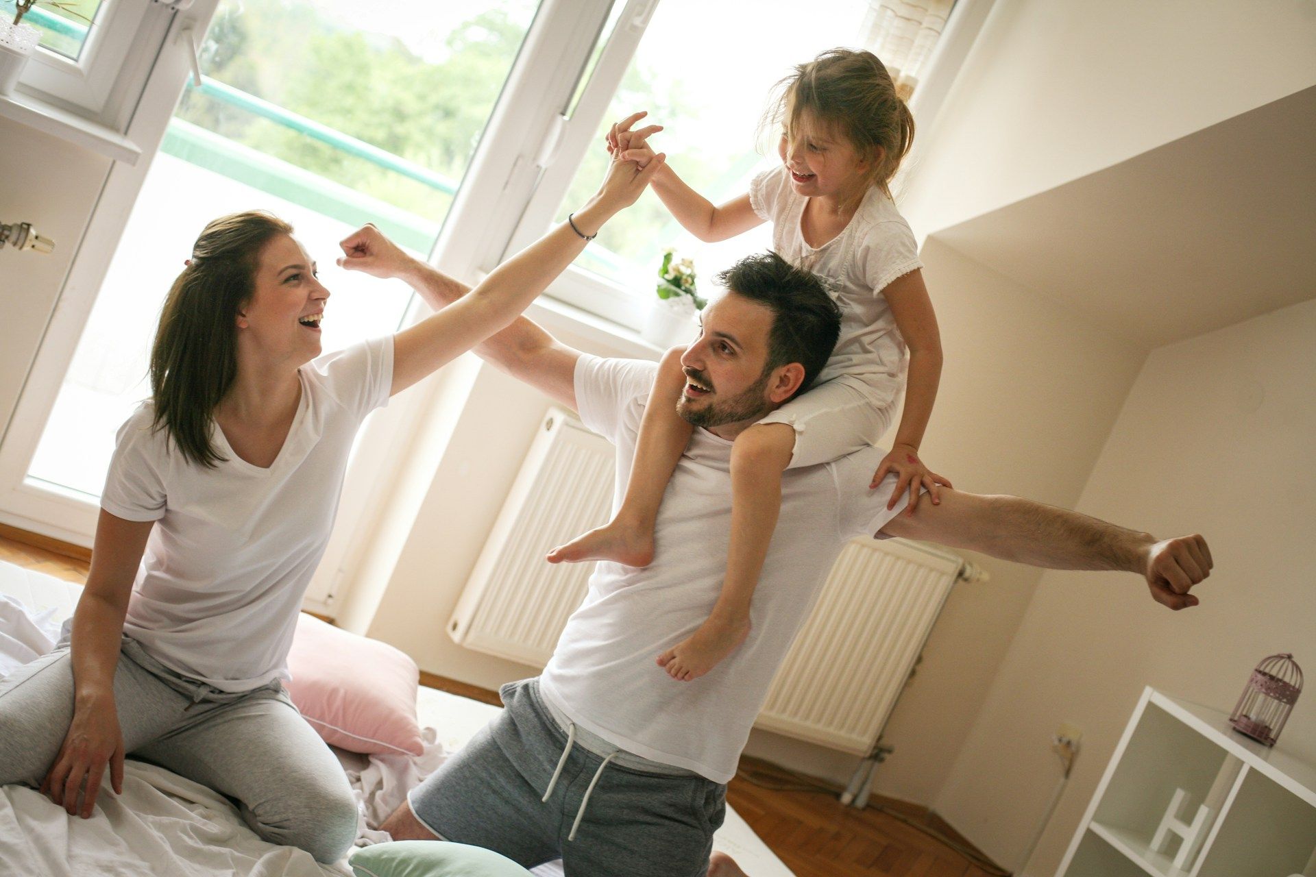 Family playing in a brightly lit bedroom: child on dad's shoulders, mom and dad raising arms, smiling.