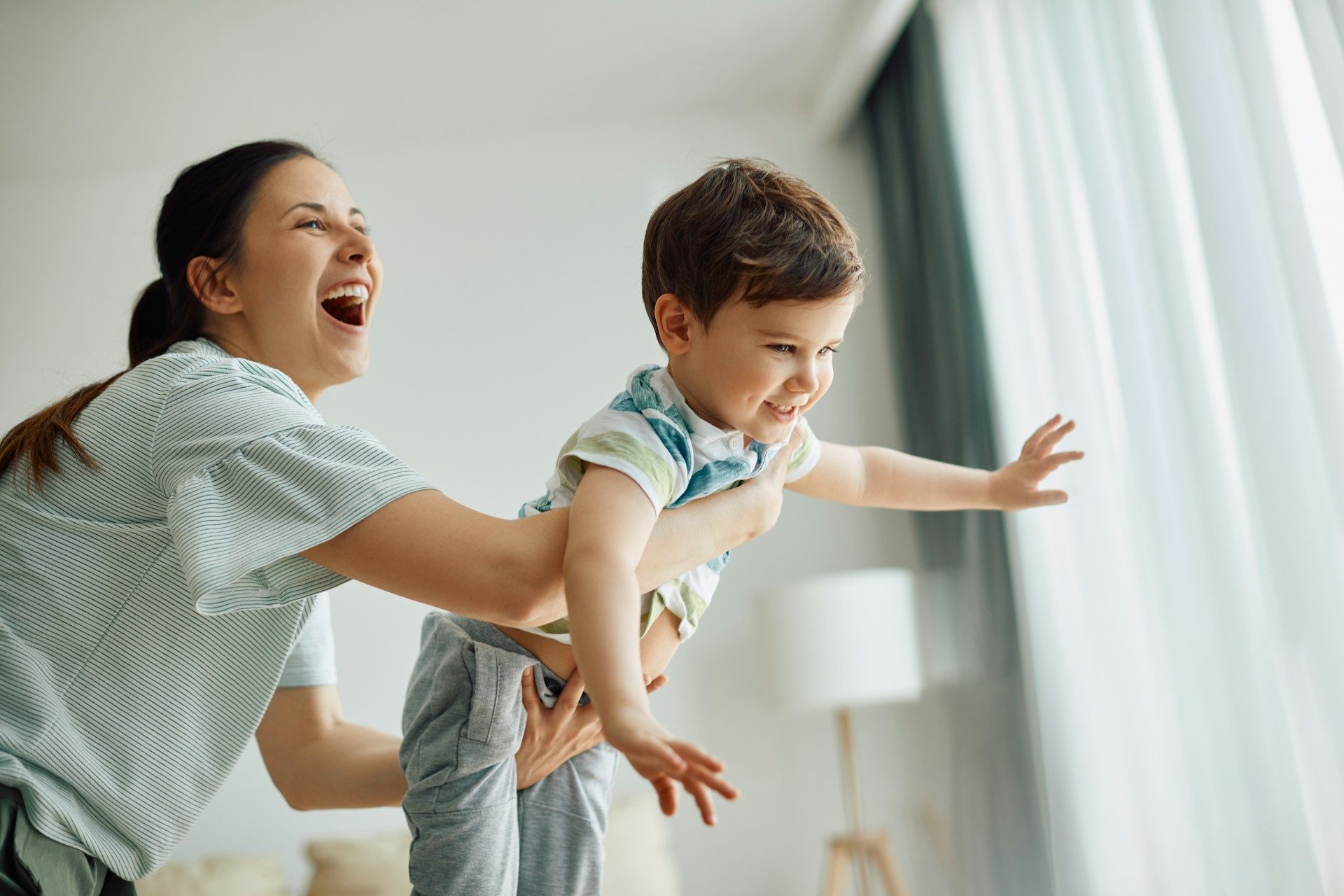 Woman lifting child, both smiling and laughing, near a window.