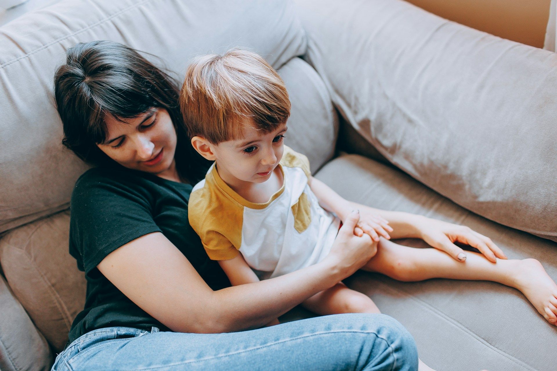 Woman and child sitting on a sofa, looking down. The child wears a yellow and white shirt.