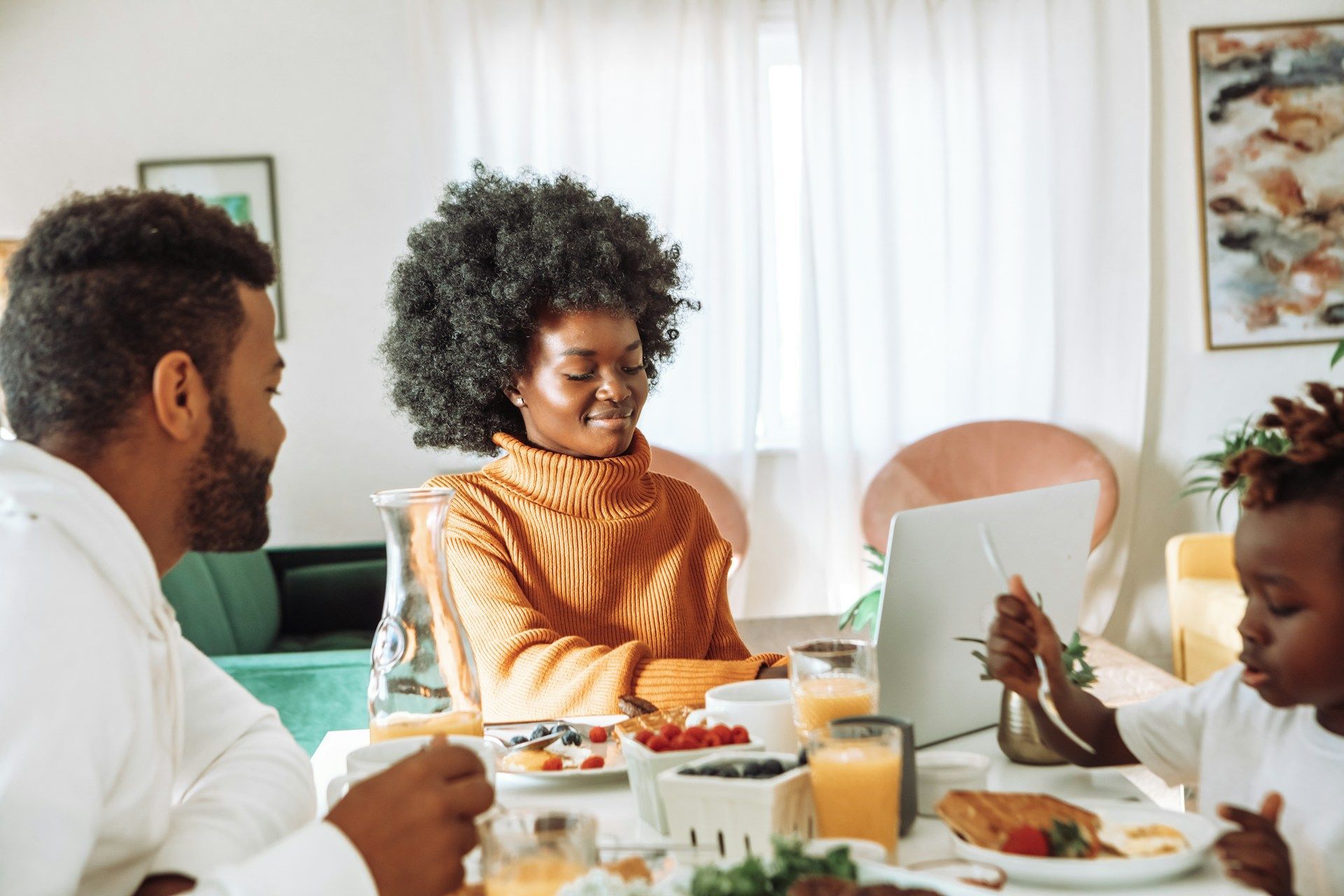 Family seated at a table eating breakfast, smiles, laptop, window in background.