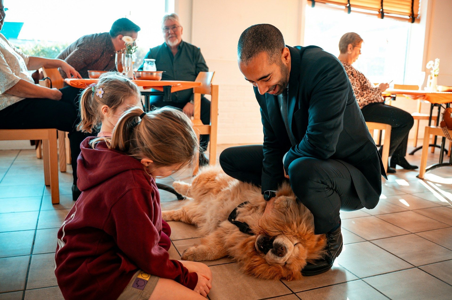 Man and child petting a large, fluffy dog inside a restaurant; other people dine at tables.