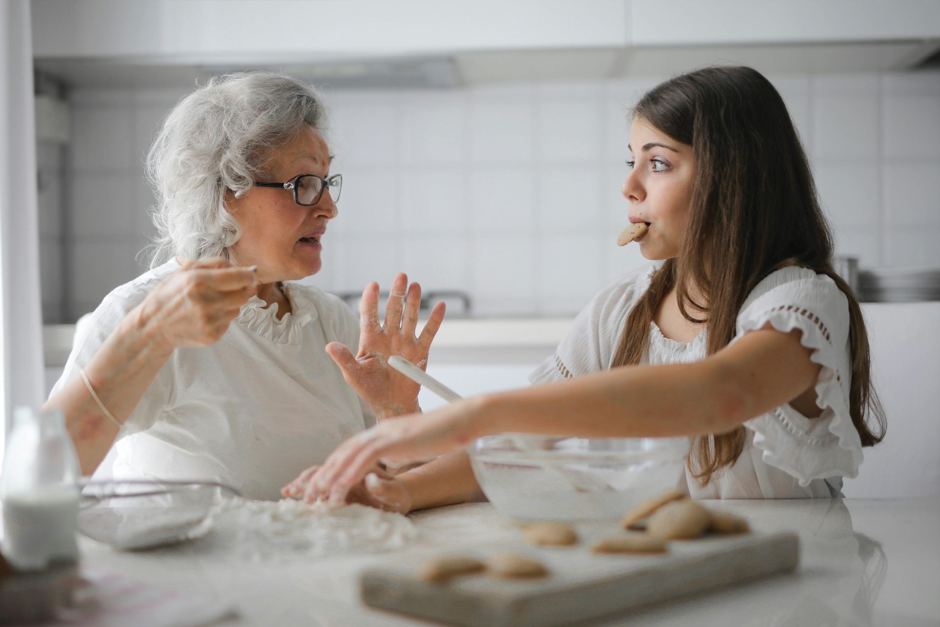 Grandmother gestures while granddaughter samples cookie dough in a bright kitchen.