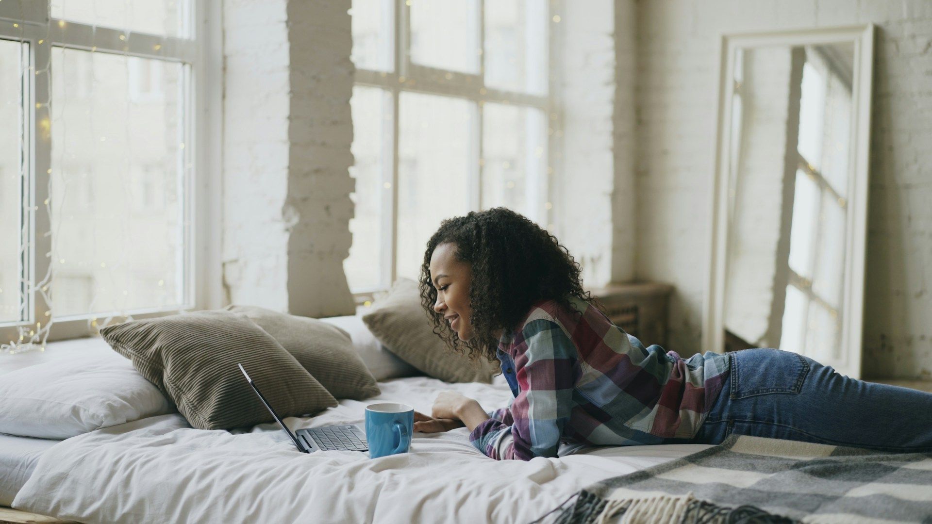 Woman lying on bed using laptop, near a window; mug nearby.