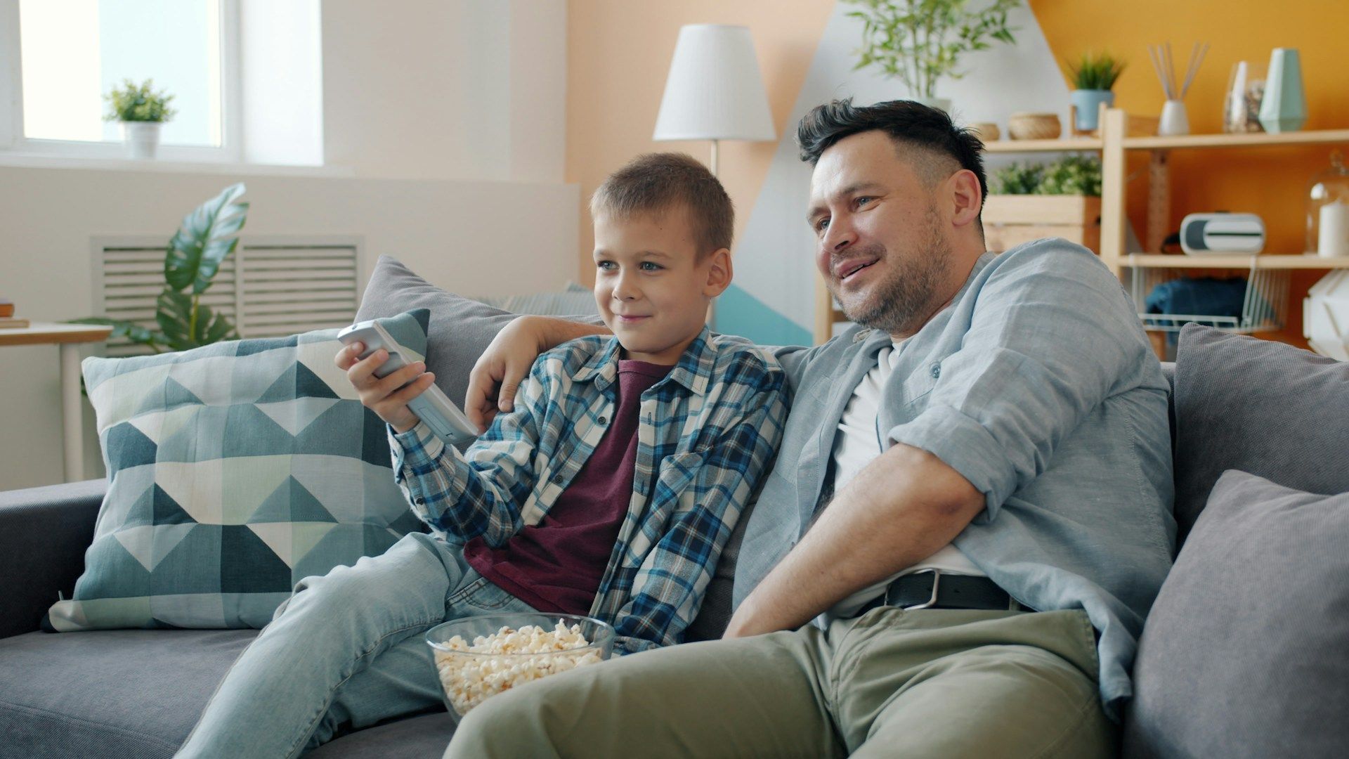 Father and son on a couch watching TV, holding remote and popcorn, smiling. Living room setting.