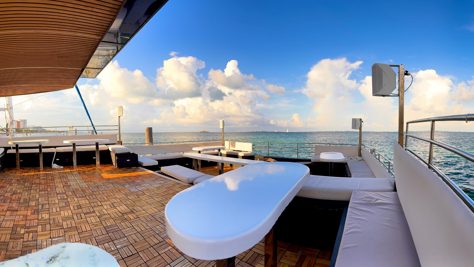 Cubierta de yate con mesas, asientos y vista al océano bajo un cielo azul con nubes hinchadas. Hix stage