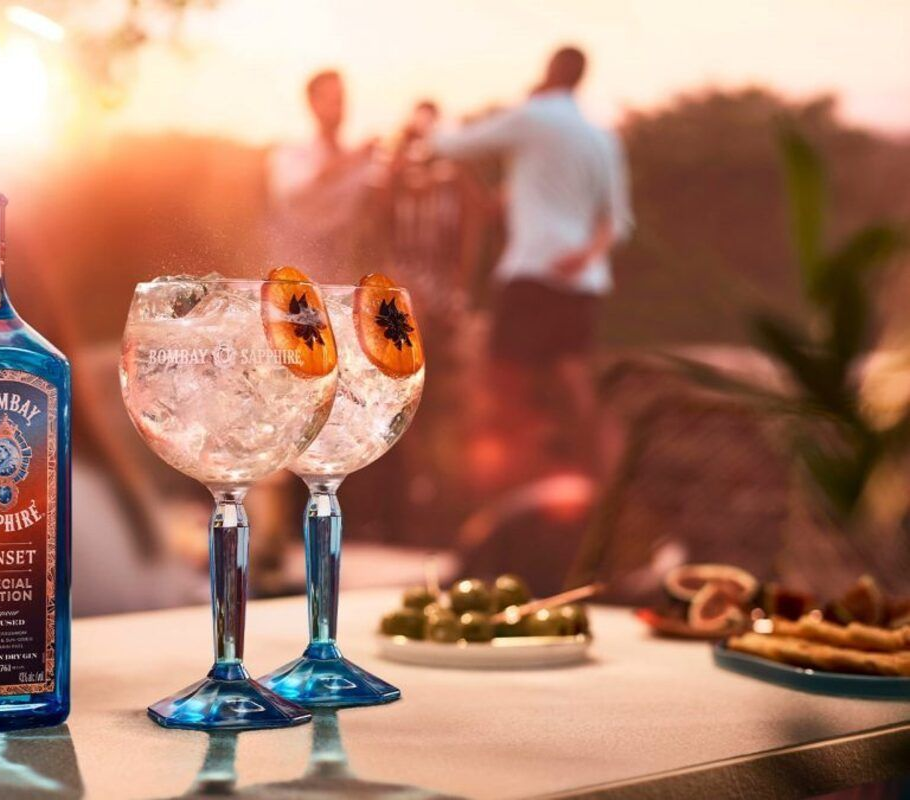 Dos cócteles de ginebra con rodajas de naranja, botella de Bombay Sapphire, entorno al aire libre con gente.