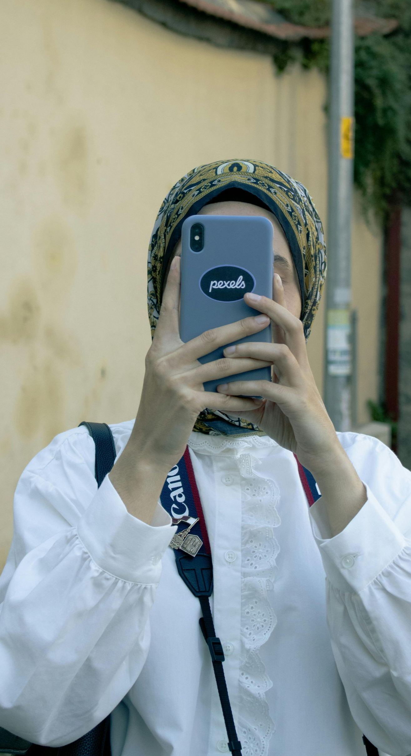Person wearing a patterned headscarf takes a photo with their phone, white shirt, street setting.