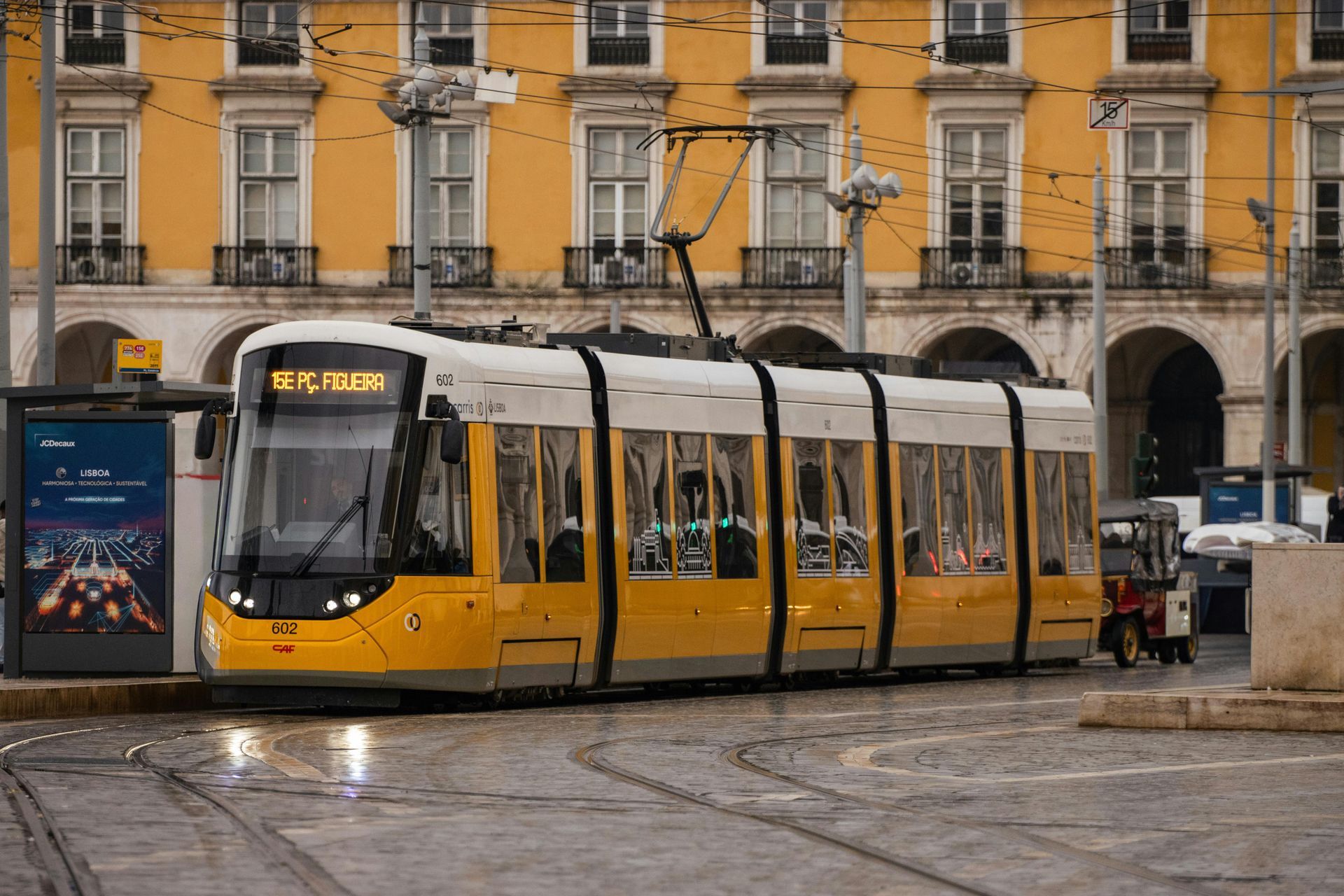 Yellow tram on tracks in front of a yellow building with many windows and an empty bus stop.