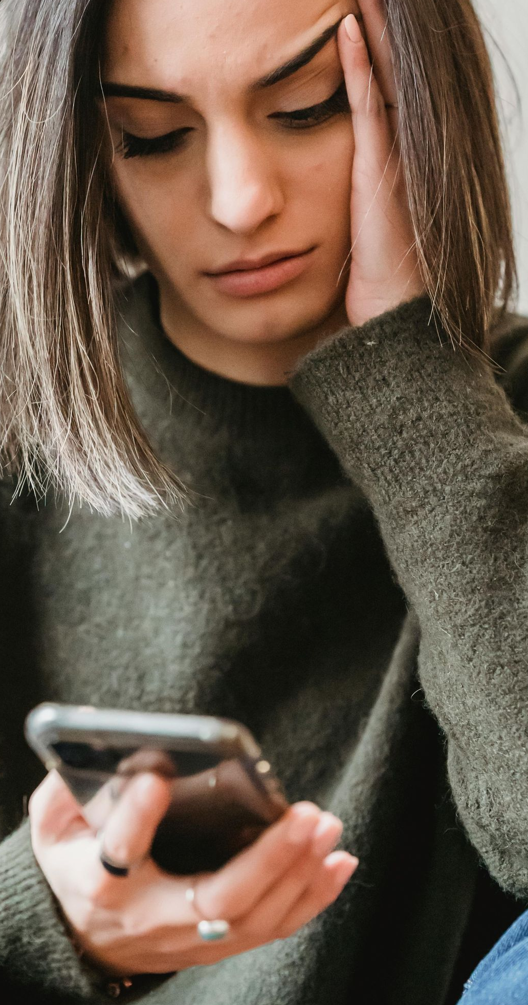 Woman with worried expression looking at phone. Olive green sweater.