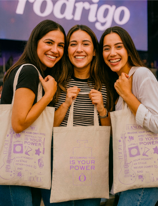 Three smiling women hold tote bags with the text 