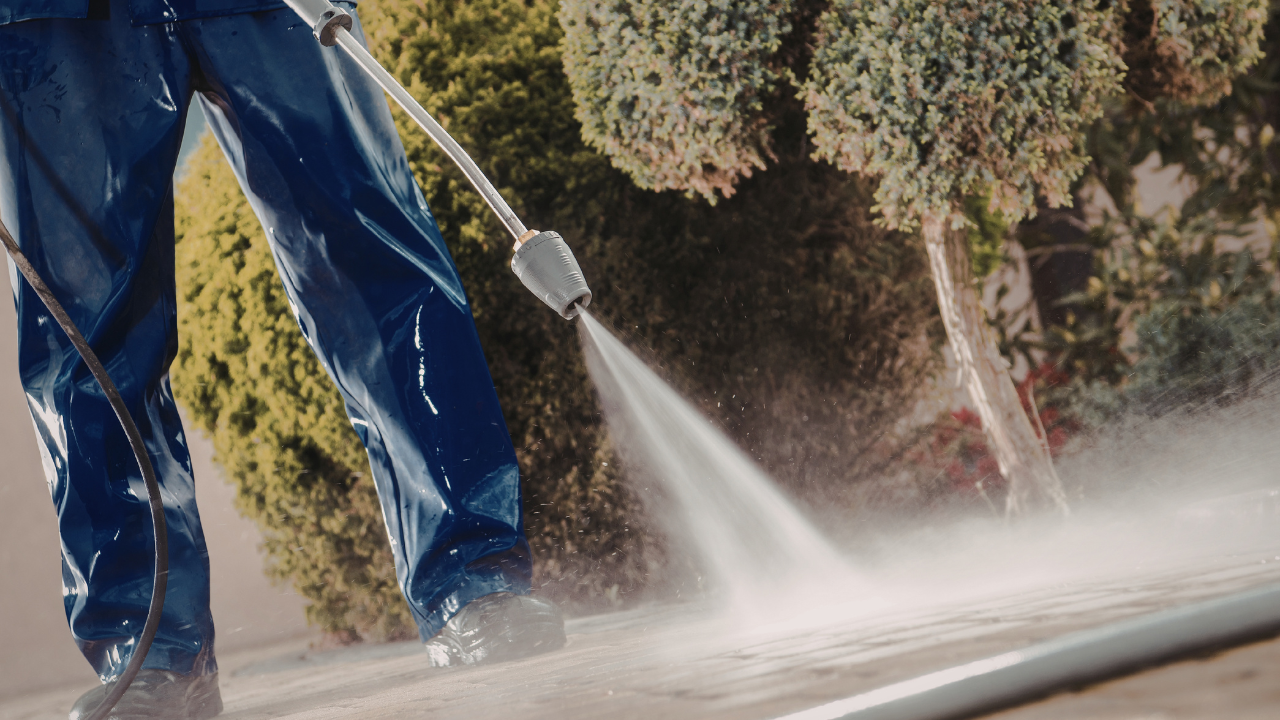 Person pressure-washing a walkway with water spraying over blue pants and nearby shrubs