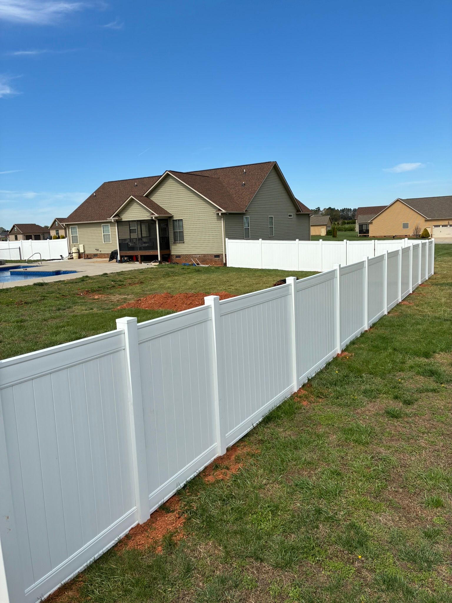 A vacant backyard with dry grass, white fence, and trees under a blue sky with clouds.