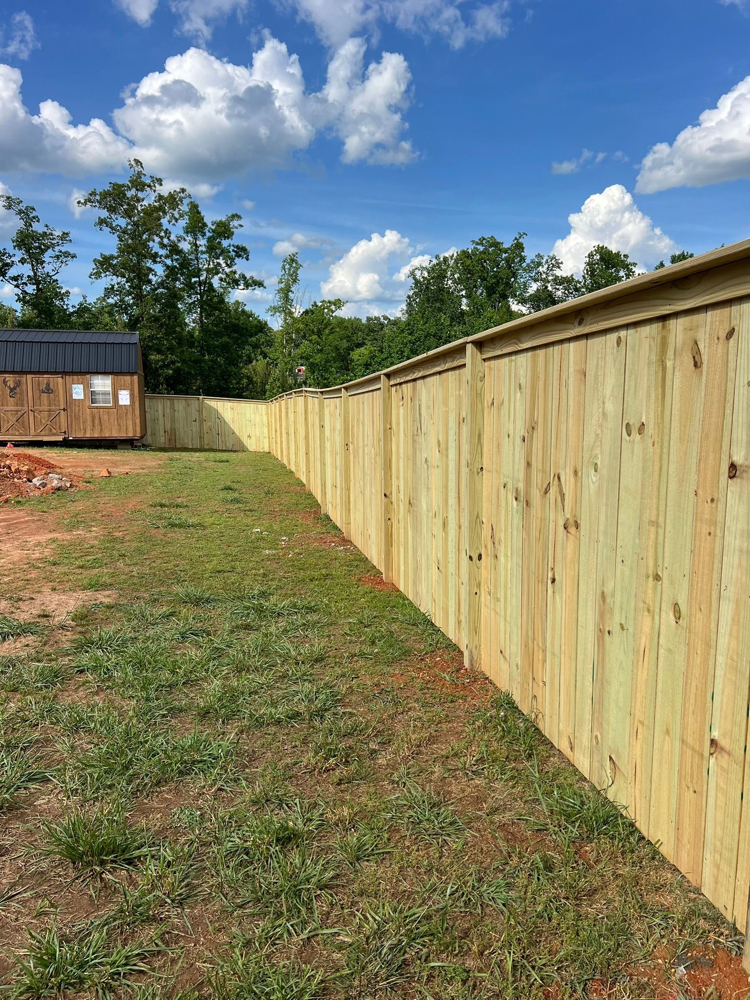 Wooden fence along a grassy yard, with a small building visible in the background against a blue sky.
