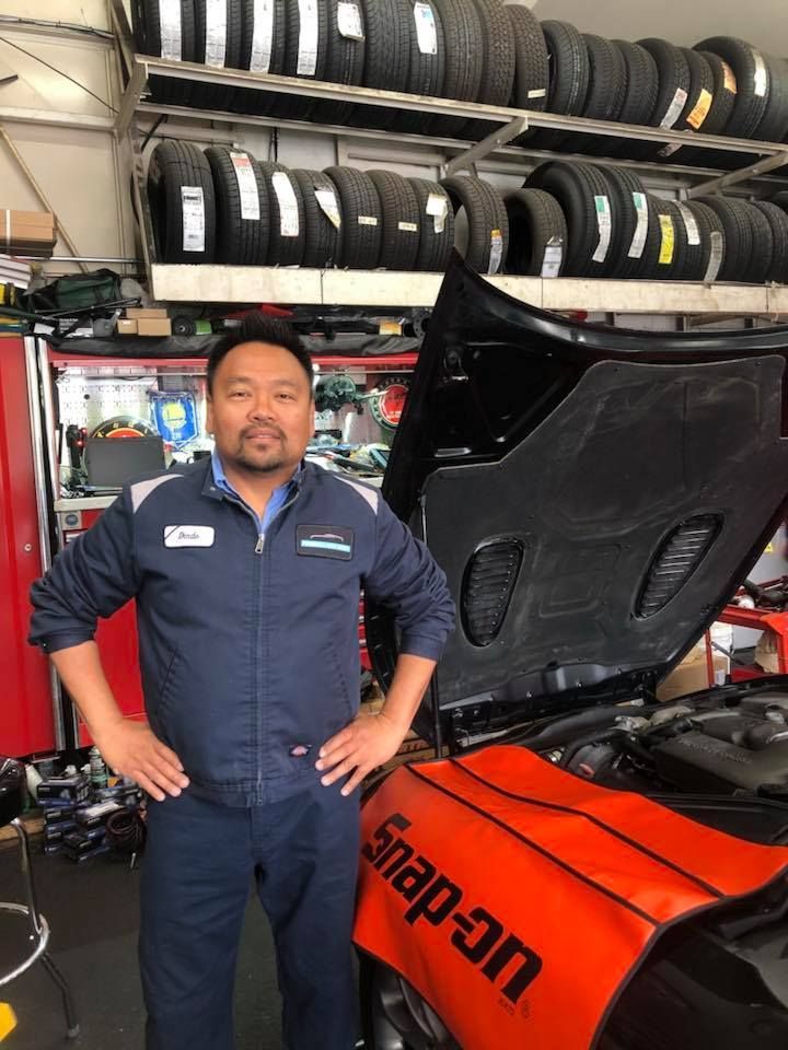 Mechanic in blue uniform stands in auto shop, hands on hips, car hood open, tires on shelf. | Pacifica Auto Works