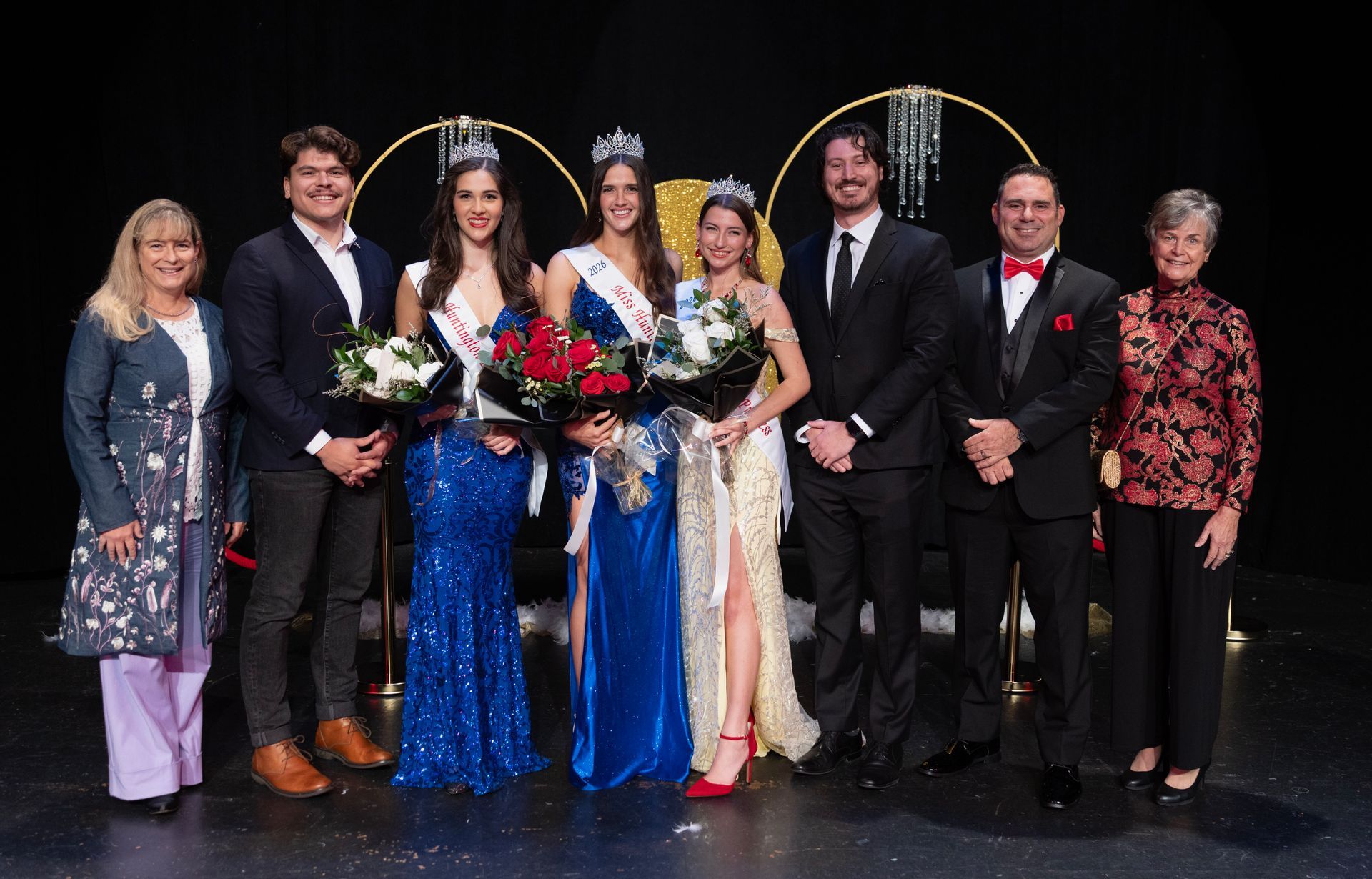 Group of people on a stage with crowns, sashes, and flowers. Black backdrop with gold circle decorations.