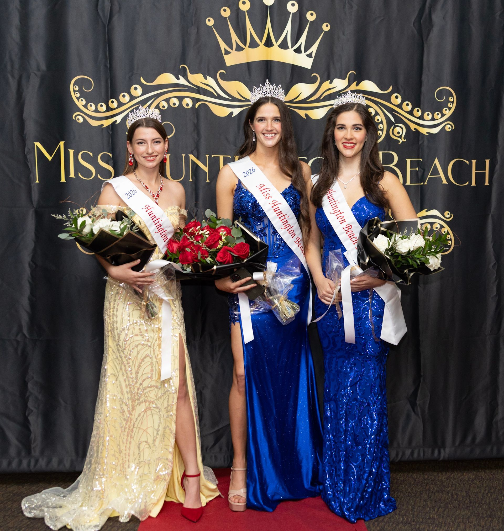 Three pageant winners in gowns, sashes, and crowns stand holding bouquets in front of a branded black backdrop.