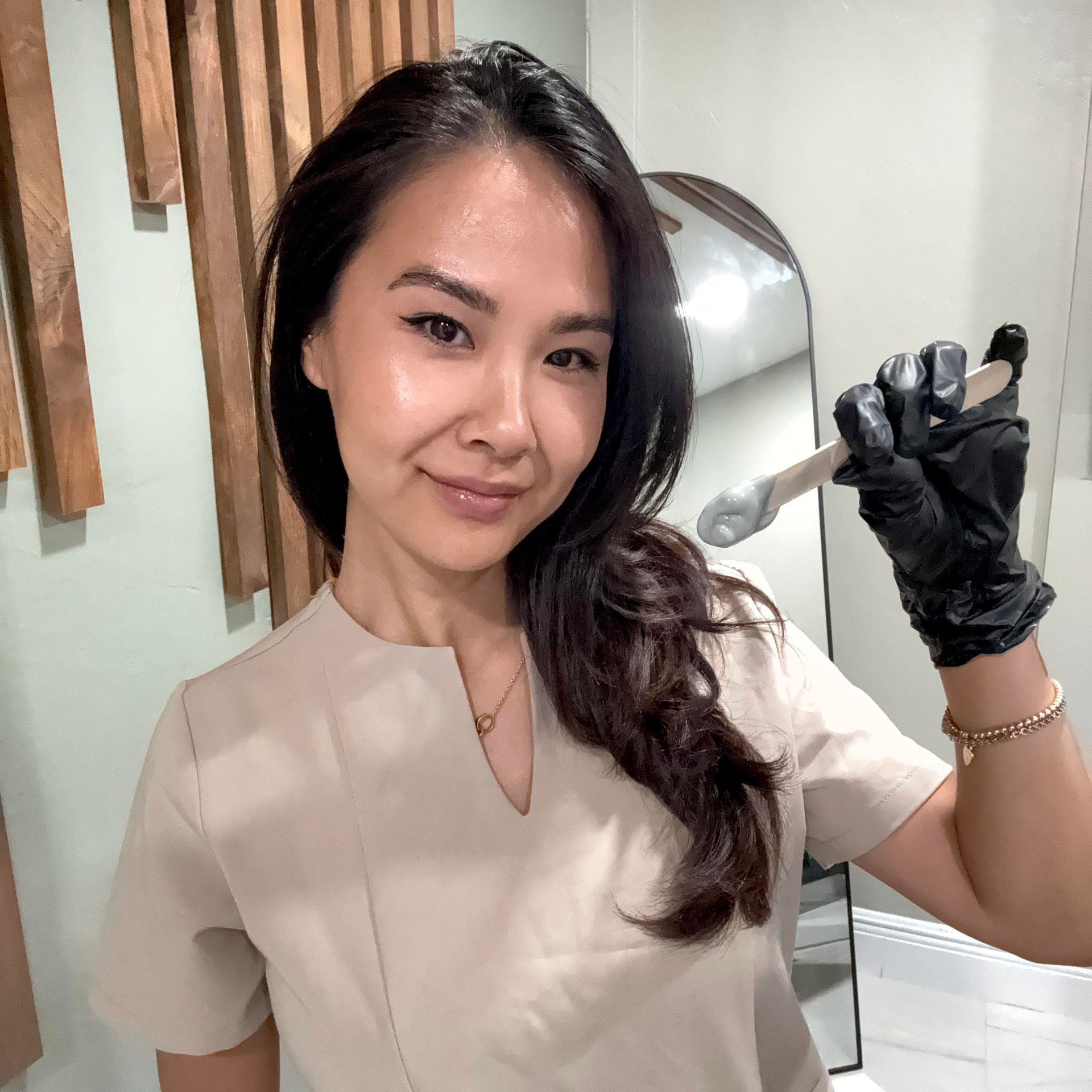Woman in tan scrubs, black glove, holding a waxing applicator, smiling indoors near a mirror.