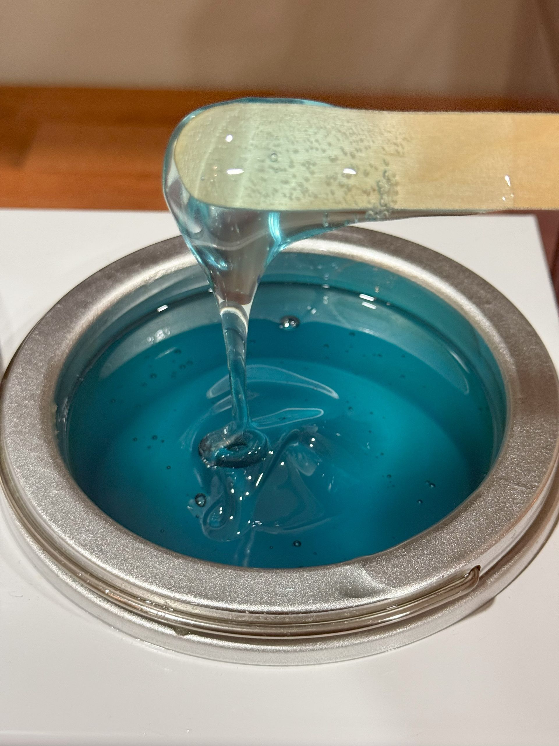 Blue wax dripping from a wooden applicator into a pot.