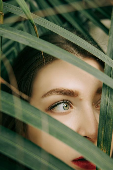 Woman's face peeking through green palm leaves, with green eyes and red lipstick, looking off-camera.