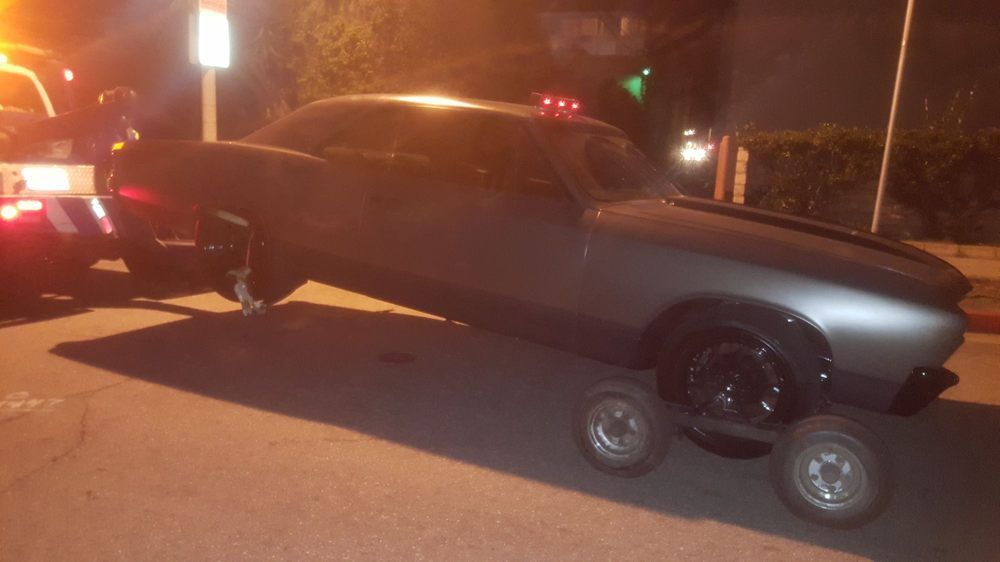 A lowrider car is being towed at night. The car is dark gray with chrome rims.
