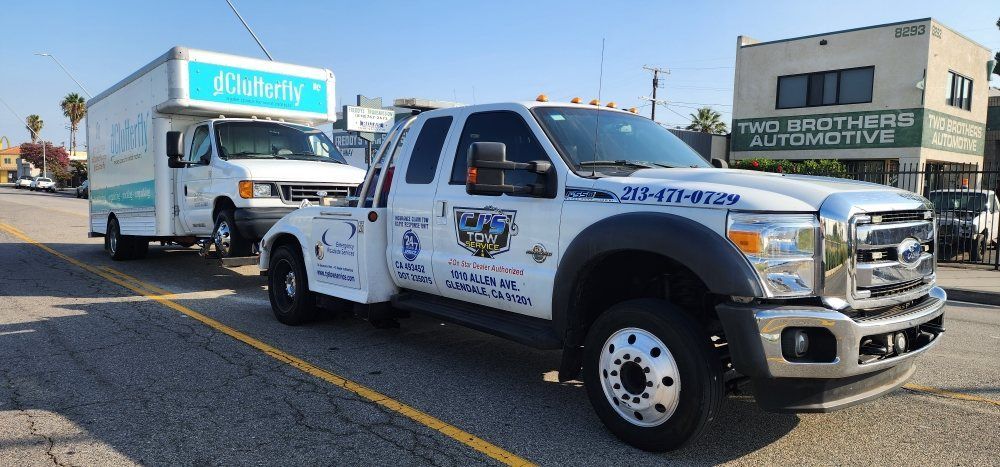 A white tow truck towing a white delivery van on a city street. Building with green sign in background.