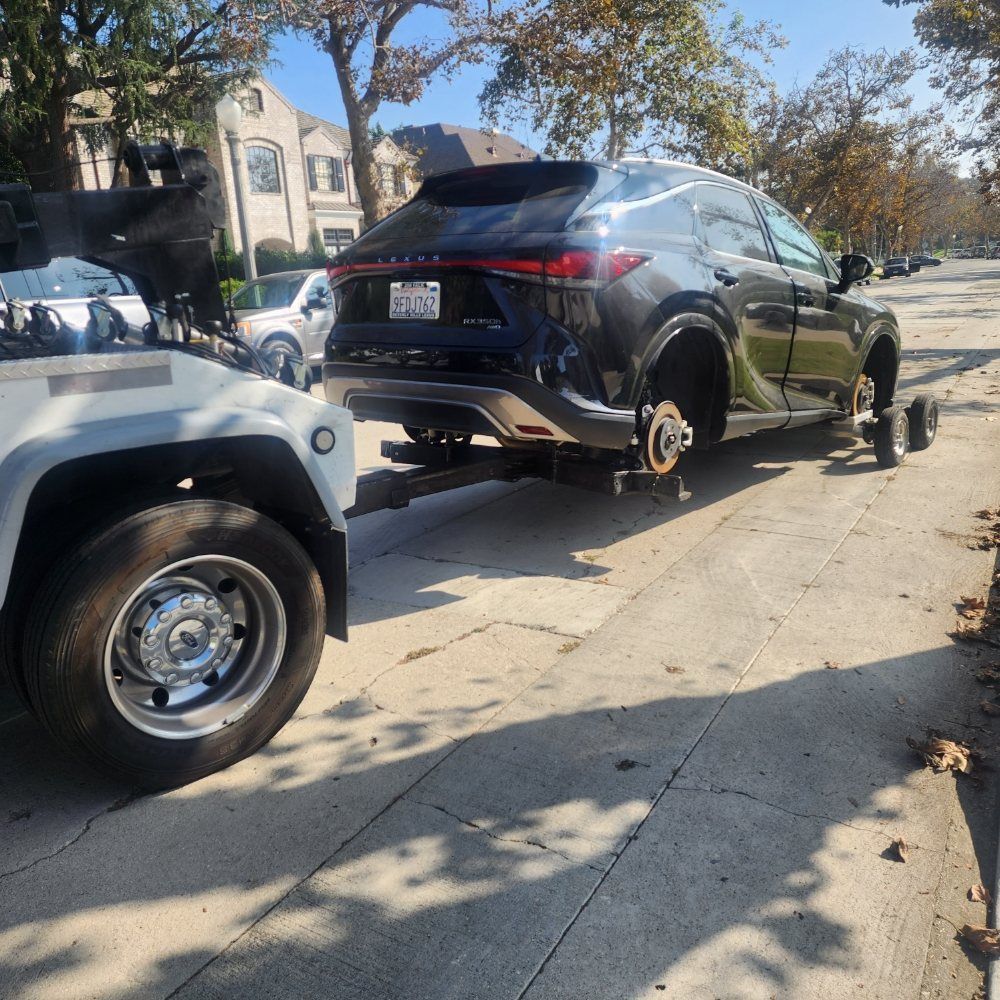 A dark SUV being towed, missing its front wheels, on a sunny street.