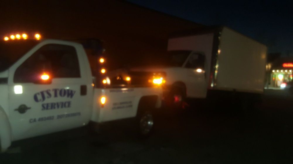 Tow truck towing a white box truck at night, lights flashing. 