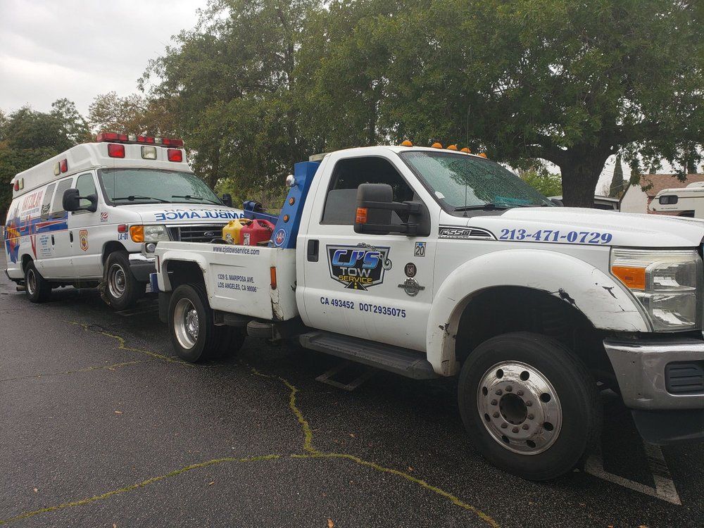 Tow truck towing an ambulance on a paved area, trees in the background. White truck and ambulance, overcast sky.