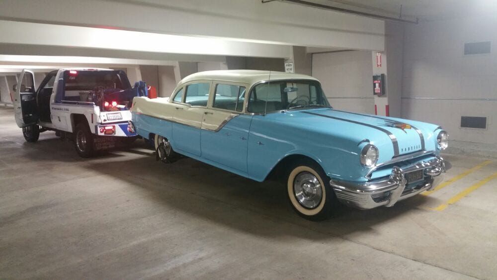 A light blue and white vintage Pontiac being towed by a tow truck in a parking garage.