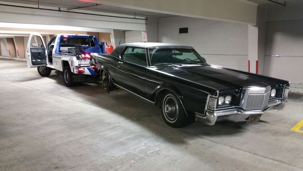 Black vintage Lincoln Continental being towed by a white tow truck in a parking garage.