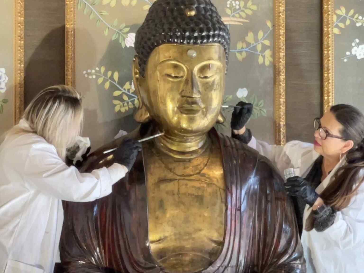 A blonde woman and brunette woman working on either side of a Buddha statue with paint brushes