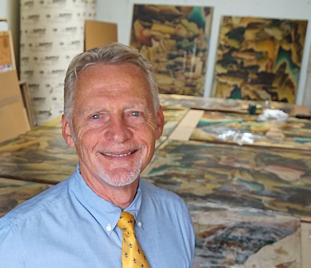 an old white man smiles while standing in front of a room full of paintings 