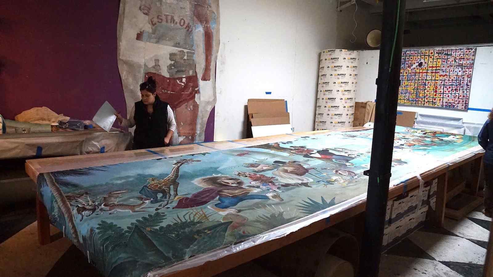 A large mural on canvas of whimsical circus animals is laid out on a long table. A woman is standing in the background with a palette.