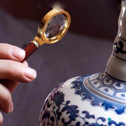 A hand with a small magnifying glass inspects a Chinese-style white and blue ceramic vase