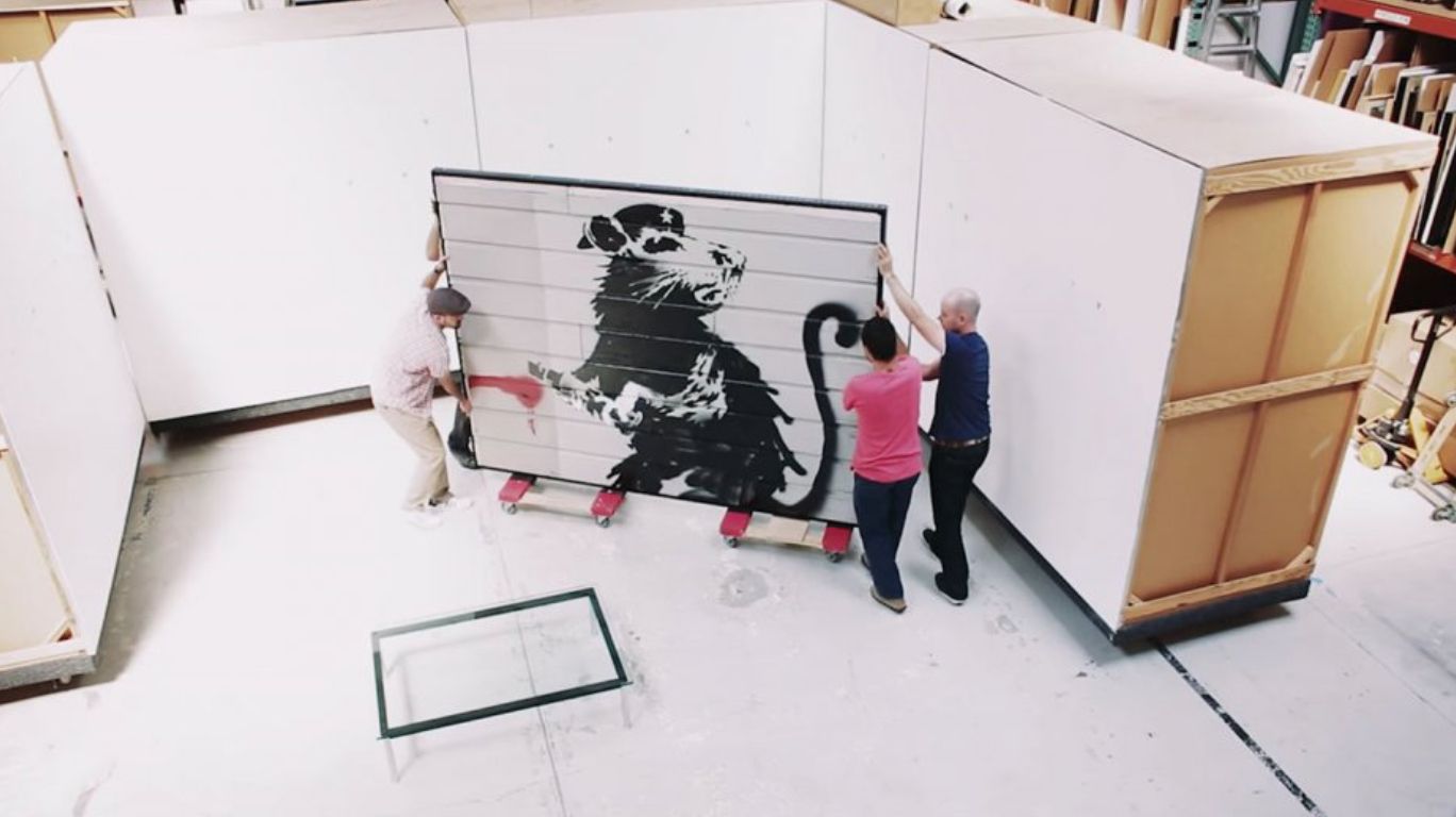 Image of 3 people in a warehouse moving a large wooden mural of a rat with a spraypaint can in its hands on two small dollies