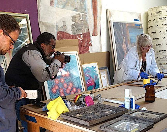 A black man with a camera photographs a woman in a lab coat working on various dirty paintings spread out on a large table