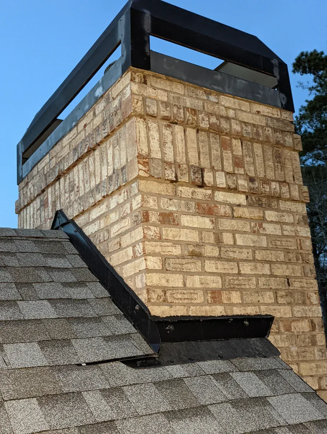 Brick chimney on a shingled roof with black metal flashing and a black cap; blue sky background.